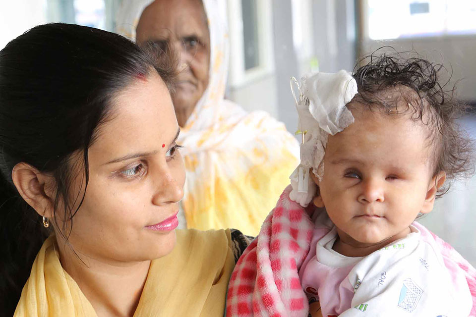 Young woman carrying a baby. The baby has bandages on his head. Behind is an older woman.